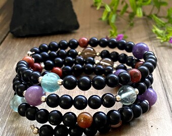 May include: A black beaded bracelet with multiple strands of smaller beads in various colors, including red, purple, blue, and green. The bracelet is coiled on a wooden surface. The text "@CrystalHealthGoddess" is visible in the lower right corner of the image.
