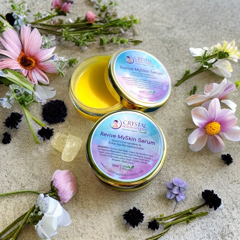 May include: A small, gold jar with a yellow cream inside. The jar is labeled "Crystal Health Goddess" and "Revive MySkin Serum". The jar is surrounded by flowers and crystals.