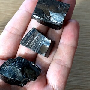 May include: Four black obsidian stones with a striated pattern. The stones are rough and have a natural, unpolished finish.