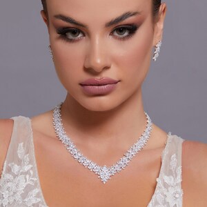 May include: A woman wearing a white lace dress with a silver necklace and earrings. The necklace is a V-shaped design with a cluster of small stones at the center.