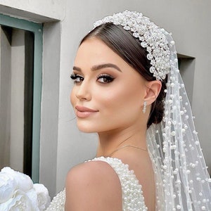 May include: A bride wearing a white beaded wedding dress and a white veil with pearl accents. The bride is wearing a silver tiara with rhinestones.