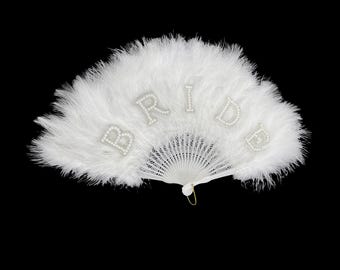 May include: A white feather fan, with the word "BRIDE" spelled out in pearl-like letters. The fan is open, showing the delicate feathers and the fan's structure. The background is black.