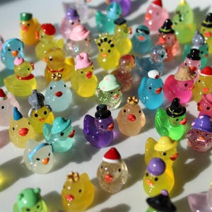 May include: A collection of miniature, colorful duck figurines. Each duck is adorned with a unique hat, including Santa hats, witch hats, and crowns. The ducks are in various colors, such as yellow, blue, pink, and purple.