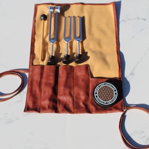 May include: A set of silver tuning forks with black bases and a wooden mallet, displayed in a brown leather roll with a tan interior. A black container with the text "Tuning Fork Activator" is also visible.