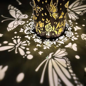 May include: A gold metal lantern with a butterfly and floral design casts a shadow of butterflies and flowers on a white surface.