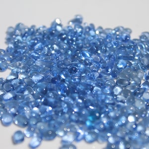 May include: A pile of small, rounded, translucent blue glass beads. The beads are scattered across a white surface, with varying shades of blue and highlights. The overall effect is a collection of decorative elements.