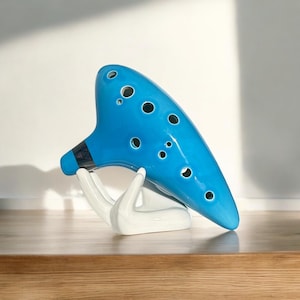 May include: A glossy, sky-blue ocarina with multiple finger holes, resting on a white ceramic hand-shaped stand. The ocarina has a triangular black and gray design near the mouthpiece. The instrument is displayed on a wooden surface.