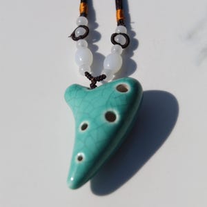 May include: A turquoise ceramic ocarina pendant with a dark crackle glaze finish and four finger holes. The instrument hangs from a dark brown cord accented with small, round, translucent white beads and orange thread details at the top.
