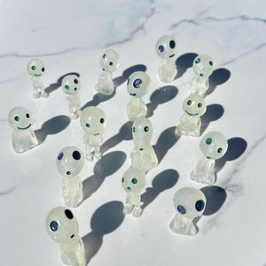 May include: A collection of small, translucent figurines with rounded heads and simple body shapes. Each figure has a unique facial expression, with eyes and mouths represented by small dots or curved lines. The figurines are arranged on a white surface.
