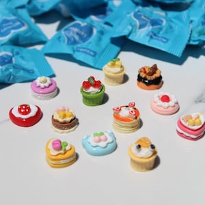 May include: A collection of miniature food-themed charms featuring various cakes, pastries, and desserts with colorful toppings like strawberries and cream, arranged in front of several blue mystery bag wrappers with white text.