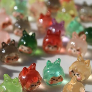 Puede incluir: Una colecci&oacute;n de amuletos transl&uacute;cidos con forma de gato de varios colores. Los amuletos son rojos, verdes, amarillos y rosas. Cada amuleto tiene una cara linda con los ojos cerrados y orejas peque&ntilde;as.