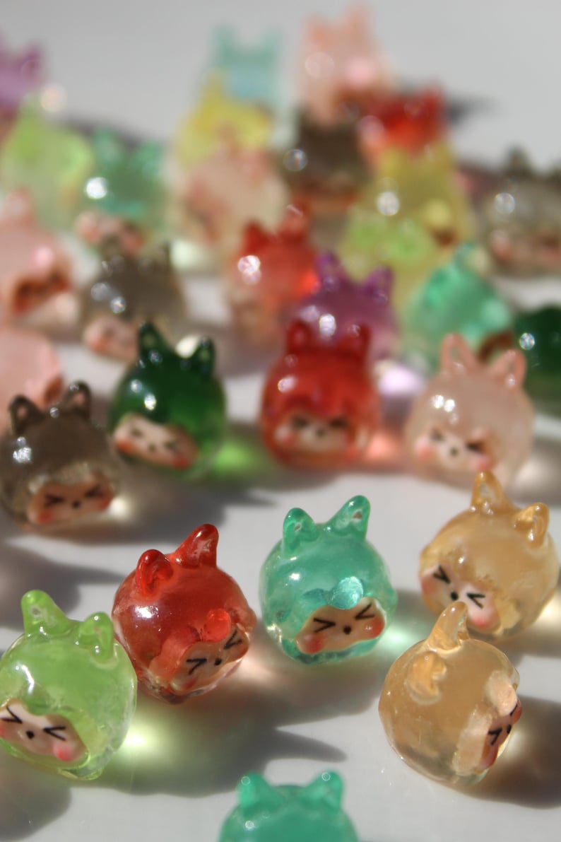 Puede incluir: Una colecci&oacute;n de amuletos transl&uacute;cidos con forma de gato de varios colores. Los amuletos son rojos, verdes, amarillos y rosas. Cada amuleto tiene una cara linda con los ojos cerrados y orejas peque&ntilde;as.