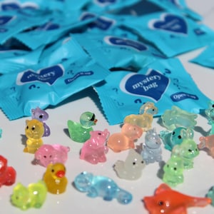 May include: A collection of colorful, translucent animal figurines, including ducks, hippos, and squirrels, are scattered in front of several blue "mystery bag" packages. The figurines are in various colors like pink, yellow, and green.