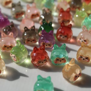 Puede incluir: Una colecci&oacute;n de peque&ntilde;as figuras transl&uacute;cidas de resina en varios colores, incluyendo rosa, verde, rojo y marr&oacute;n. Cada figura tiene una cara de dibujos animados y un sombrero o capucha, creando una est&eacute;tica caprichosa. Las figuras est&aacute;n dispersas sobre una superficie blanca.