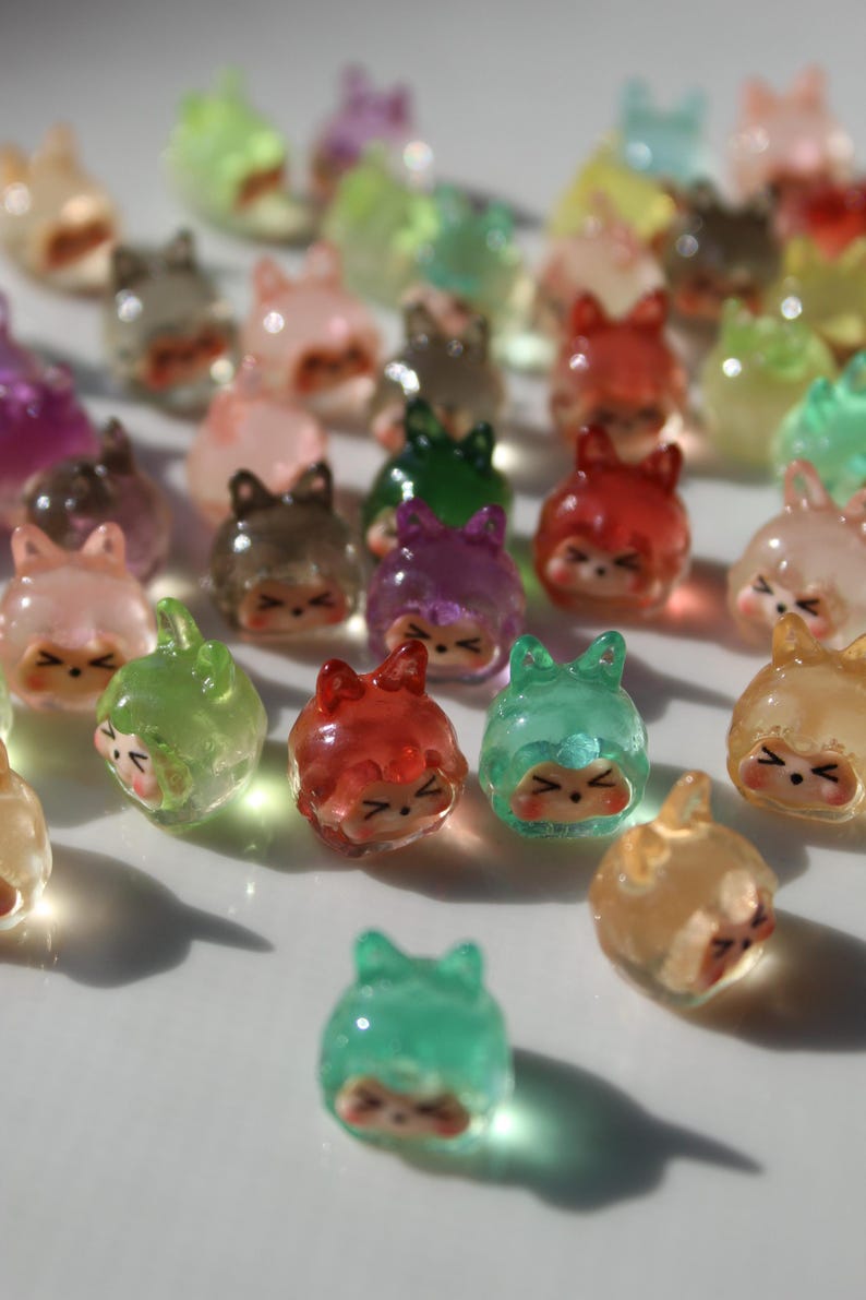 Puede incluir: Una colecci&oacute;n de peque&ntilde;as figuras transl&uacute;cidas de resina en varios colores, incluyendo rosa, verde, rojo y marr&oacute;n. Cada figura tiene una cara de dibujos animados y un sombrero o capucha, creando una est&eacute;tica caprichosa. Las figuras est&aacute;n dispersas sobre una superficie blanca.