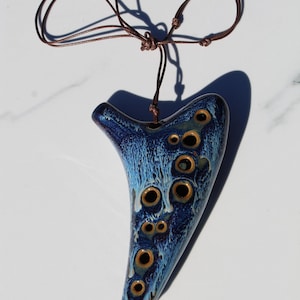 May include: A ceramic ocarina in a teardrop shape, with a dark blue glaze and numerous circular holes. The instrument has a brown cord for wearing around the neck. The ocarina's surface has a textured, mottled appearance.