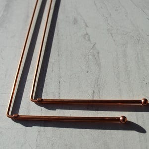 May include: Two copper-colored dowsing rods, each with a ball-shaped end. The rods are bent at a right angle, with one longer than the other. The rods are lying on a light gray surface, casting shadows.