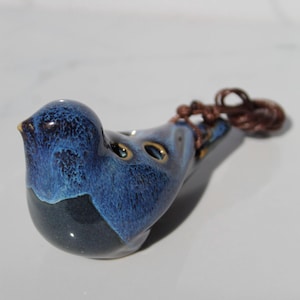 Puede incluir: Un silbato de cerámica azul y negro en forma de pájaro con un cordón marrón. El silbato tiene un esmalte moteado y cuatro agujeros para los dedos.