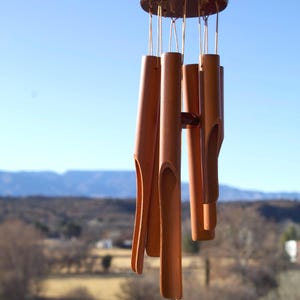 May include: A wooden wind chime with a dark brown top and cylindrical chimes. The chimes are a light brown color and have a curved design. The wind chime hangs from a thin, beige string against a blue sky.