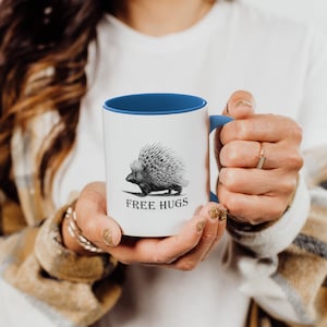 Puede incluir: Una taza de cerámica blanca con un borde y un asa azules. La taza presenta una ilustración en blanco y negro de un puercoespín con el texto "FREE HUGS" debajo.