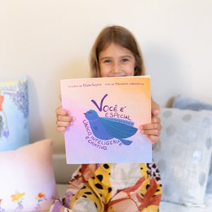 May include: A children's book with a colourful cover featuring a blue bird illustration and the words "Você é Especial" in purple. The book is held by a person, with decorative pillows in the background.