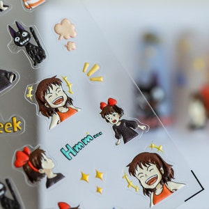 May include: A sheet of clear stickers featuring cartoon characters from the animated film 'Howl's Moving Castle'. The stickers include a black cat, a girl with red hair and a red bow, and a girl with brown hair and a red bow. The stickers also include text that says 'Hmm...' and 'eek'.