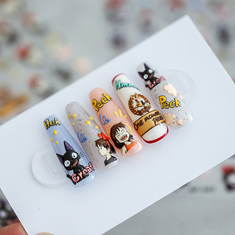 May include: Five long, light blue, pink, and white artificial nails with a black cat, a girl with brown hair, and a cartoon character with a red bow on her head. The nails have glitter and the words "Heh Great", "Peek", "One moment please", and "Peek" written on them.