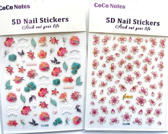May include: Two packs of 5D nail stickers from CoCo Notes. One pack features colorful floral designs with leaves and pearls. The other pack has pink cherry blossom designs with gold accents. The text "5D Nail Stickers" and "Stick out your life" is visible.