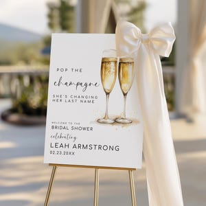 Puede incluir: Un letrero blanco sobre un caballete dorado con el texto "Pop the champagne" y "Bienvenida a la despedida de soltera que celebra a Leah Armstrong". Se ilustran dos copas de champán. Un lazo blanco y tela cubren el lateral.