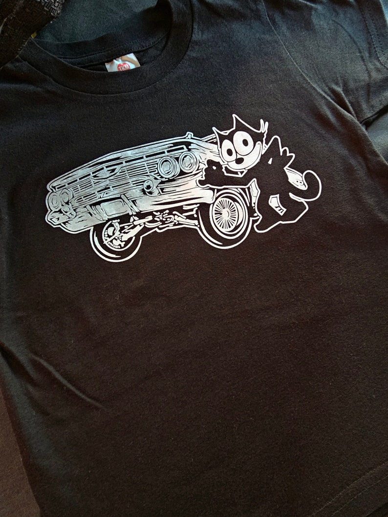 Może przedstawiać: Czarny T-shirt z białym nadrukiem przedstawiającym kresk&oacute;wkowego kota prowadzącego klasyczny samoch&oacute;d.