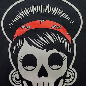 May include: A black and white illustration of a skull with a red bandana and a halo. The skull has large eyes and a small nose. The bandana has a paisley pattern.