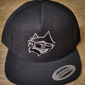 Puede incluir: Gorra de béisbol negra con un gráfico bordado en blanco y negro de un gato con gafas de sol. La gorra tiene una etiqueta blanca que dice "The Authentic Yupoong Snapback Yupoong Since 1974".