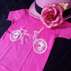 May include: A pink t-shirt with a white graphic of a bicycle on it. The shirt is laying on a black surface with a pink hat with a rose on it.