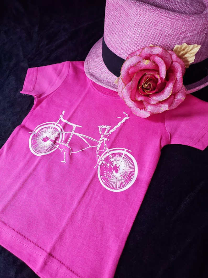 May include: A pink t-shirt with a white graphic of a bicycle on it. The shirt is laying on a black surface with a pink hat with a rose on it.