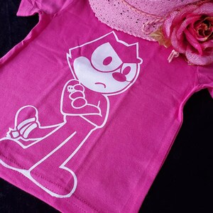 May include: A pink t-shirt with a white cartoon character wearing a hat and holding a small object. The character has a large head and a small body. The shirt has a short sleeve and a crew neck.