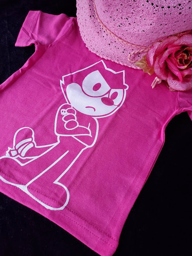 May include: A pink t-shirt with a white cartoon character wearing a hat and holding a small object. The character has a large head and a small body. The shirt has a short sleeve and a crew neck.