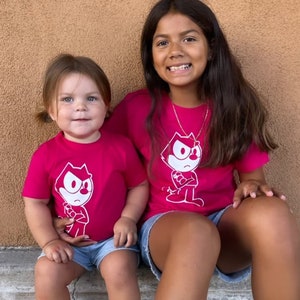 May include: Two girls wearing pink t-shirts with a white cartoon cat graphic. The cat is wearing a white hat and has a mischievous expression. The girls are sitting on a step with a brown background.