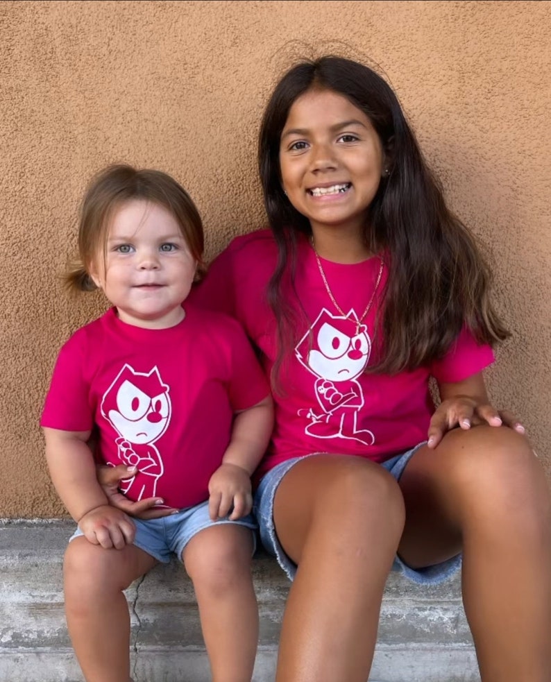 May include: Two girls wearing pink t-shirts with a white cartoon cat graphic. The cat is wearing a white hat and has a mischievous expression. The girls are sitting on a step with a brown background.