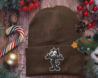 Puede incluir: Un gorro de punto marrón con un gato de dibujos animados en blanco y negro bordado en la parte delantera. El gorro está rodeado de decoraciones navideñas, como un bastón de caramelo, una rama de pino y bayas rojas.