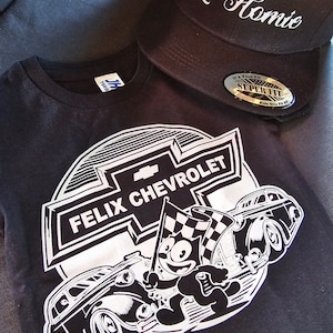 Puede incluir: Camiseta negra con un gráfico blanco de Félix el Gato conduciendo un Chevrolet vintage con el texto "Felix Chevrolet" sobre el coche. La camiseta está sobre una superficie azul con una gorra de béisbol negra que dice "Lil Homie" en cursiva blanca.