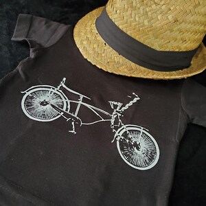 May include: A black t-shirt with a white graphic of a bicycle. The t-shirt is lying on a black surface with a straw hat on top of it.