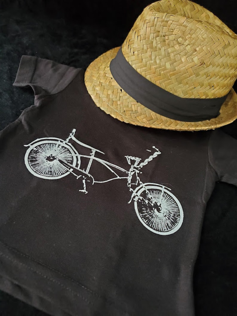 May include: A black t-shirt with a white graphic of a bicycle. The t-shirt is lying on a black surface with a straw hat on top of it.