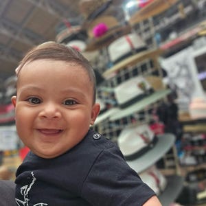 May include: A close-up of a smiling baby wearing a black t-shirt with a white bicycle design. The baby is in focus, while the background is blurred and shows a variety of hats stacked on shelves.