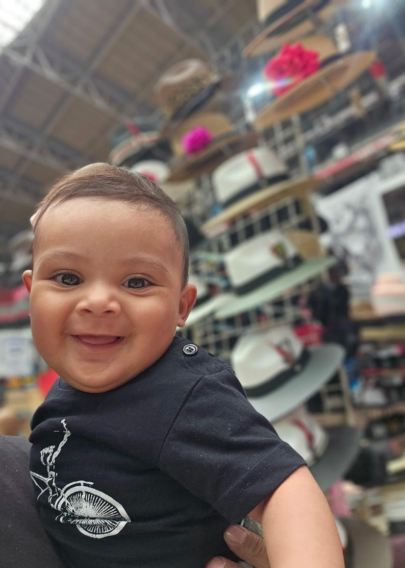 May include: A close-up of a smiling baby wearing a black t-shirt with a white bicycle design. The baby is in focus, while the background is blurred and shows a variety of hats stacked on shelves.
