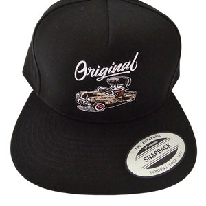 Puede incluir: Gorra de béisbol negra con un logotipo "Original" bordado en blanco y un diseño de coche de dibujos animados. La gorra tiene una visera negra y una etiqueta blanca "The Authentic Yupoong Snapback" en la parte posterior.