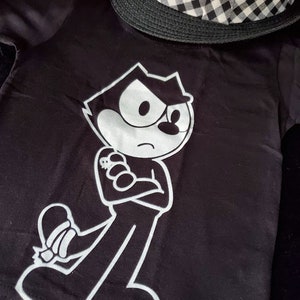 May include: Black t-shirt with a white graphic of a cartoon cat with a mischievous expression. The cat is wearing a white shirt and has a white outline.