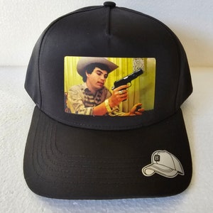 May include: Black baseball cap with a white patch on the side that says "B". The front of the cap features a square image of a man wearing a cowboy hat holding a gun.