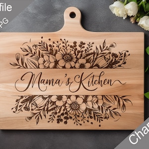 May include: A wooden cutting board with a floral design and the text "Mama's Kitchen" engraved on it. The board is light brown and has a handle on the top. The board is on a gray surface.