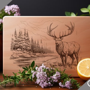Peut inclure: Une planche à découper en bois avec un motif gravé au laser représentant un cerf debout dans une forêt au bord d'un lac. Le texte "Charcuterie design" est dans le coin inférieur droit de la planche.
