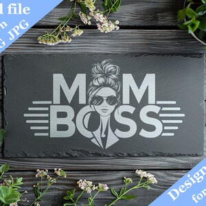 May include: A black slate sign with the text "MOM BOSS" in silver lettering. A stylized image of a woman with a bun and sunglasses is in the center of the text. The sign is on a wooden surface with green plants and white flowers.